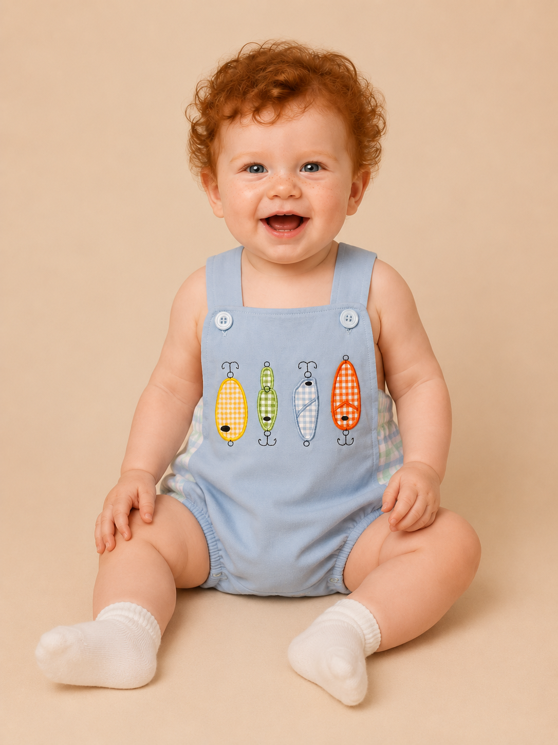 A smiling baby with curly red hair sits on a beige studio background wearing a light blue sleeveless bubble with four colorful fish graphics on the front and white socks.