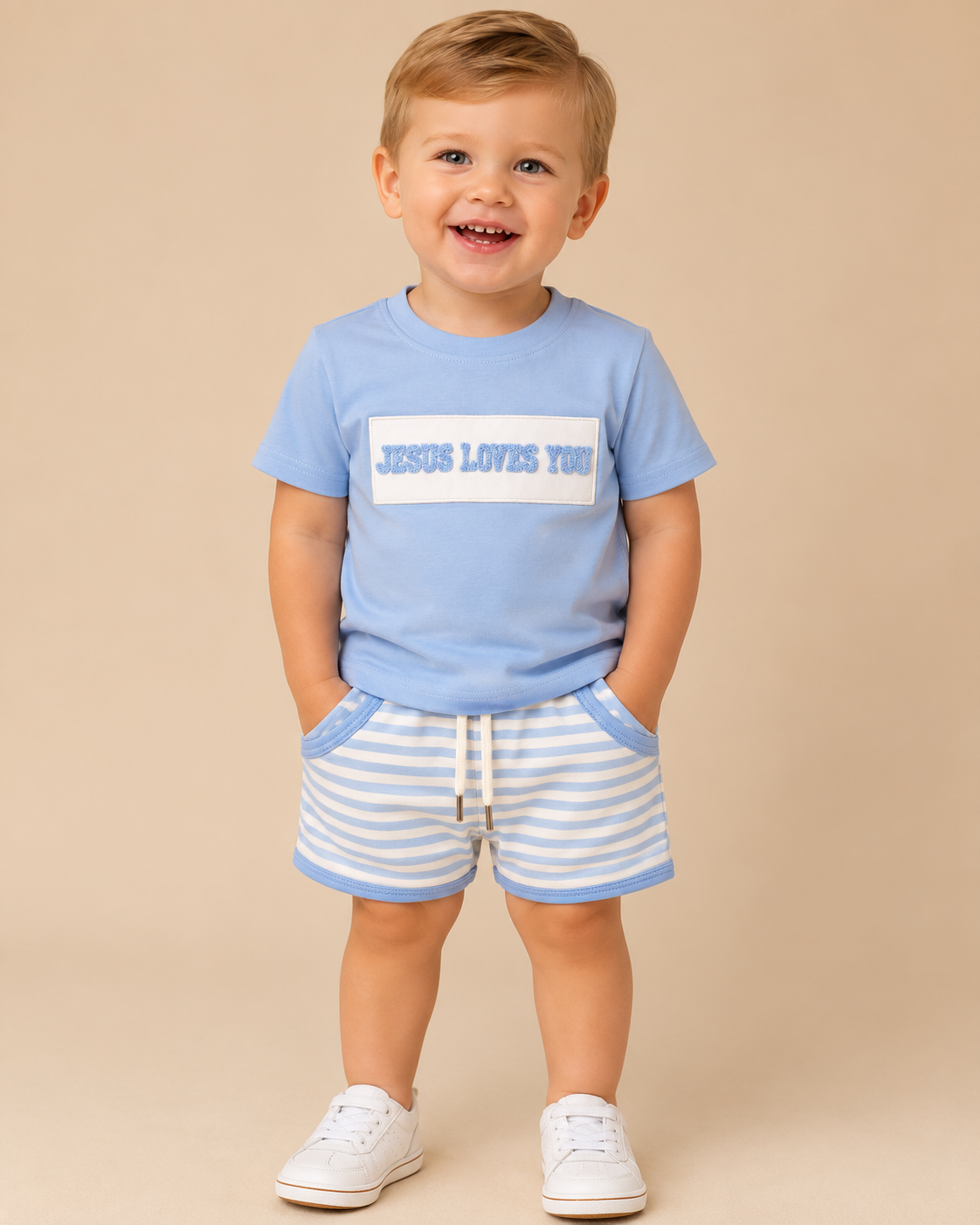 A toddler standing against a solid beige background, wearing a light blue t-shirt with a white graphic box and the text 'JESUS LOVES YOU' in blue, paired with light blue and white striped shorts and white sneakers.