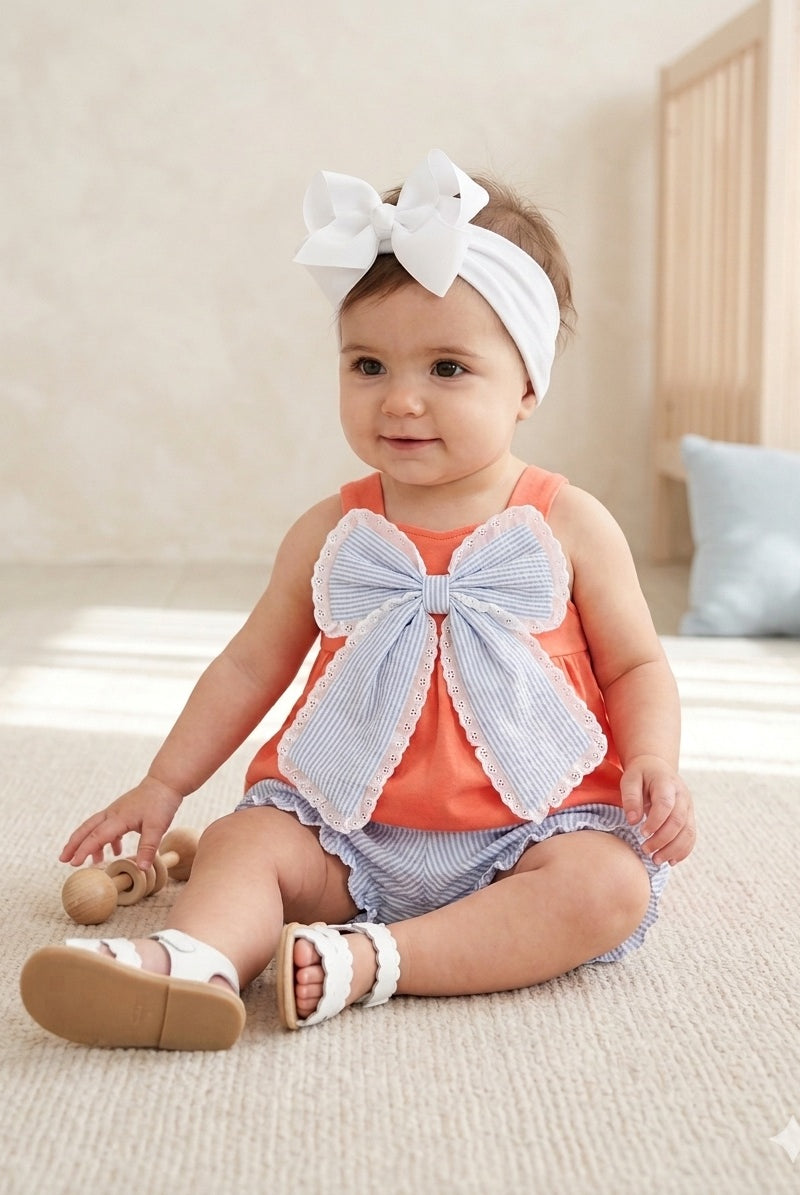 Baby wearing an orange outfit with a large bow sitting on a carpeted floor.