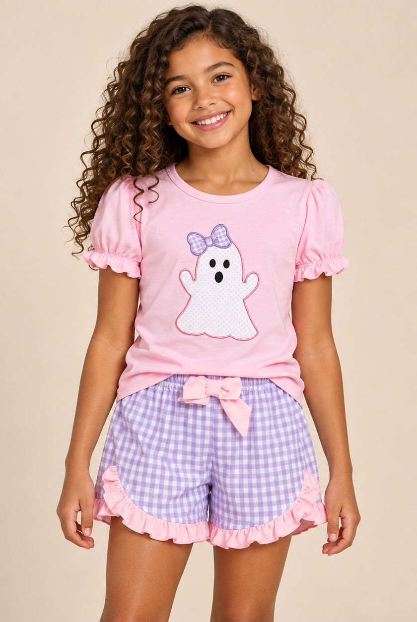 A young girl with curly brown hair smiling, wearing a pink short-sleeve pajama top with a white ghost graphic and purple bow, paired with lavender and white gingham shorts with pink ruffle trim.
