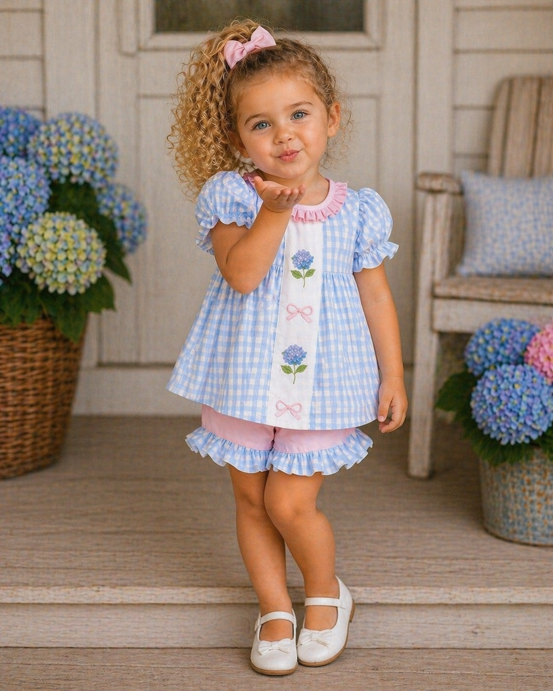 Young girl in a blue checkered dress with floral design, standing on a porch with hydrangeas in the background.