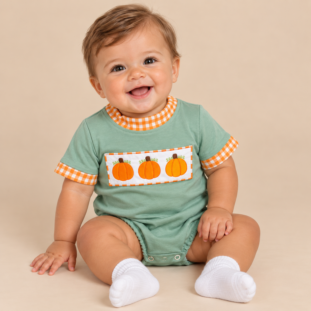 A smiling baby sitting on a beige studio background, wearing a sage green short-sleeve romper with orange and white gingham trim, a chest patch with three orange pumpkins, and white socks.