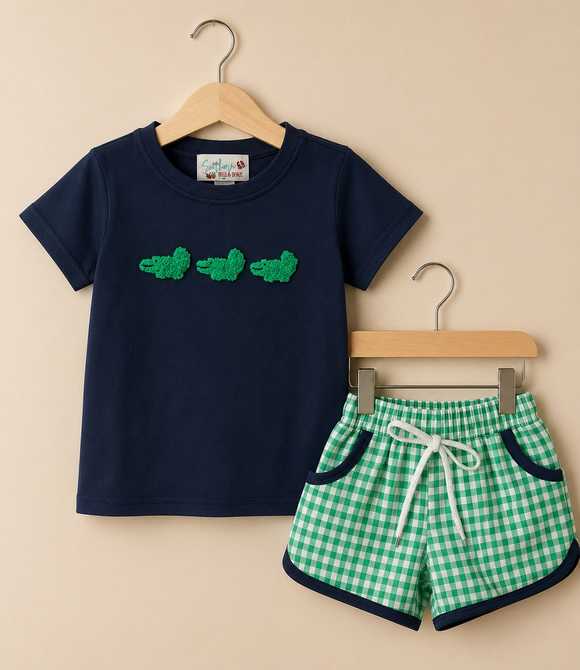 A children's two-piece outfit set consisting of a navy blue short-sleeve t-shirt with green crocodile appliques on the chest, and green and white gingham check shorts with navy blue trim and a white drawstring, displayed on wooden hangers against a beige background.