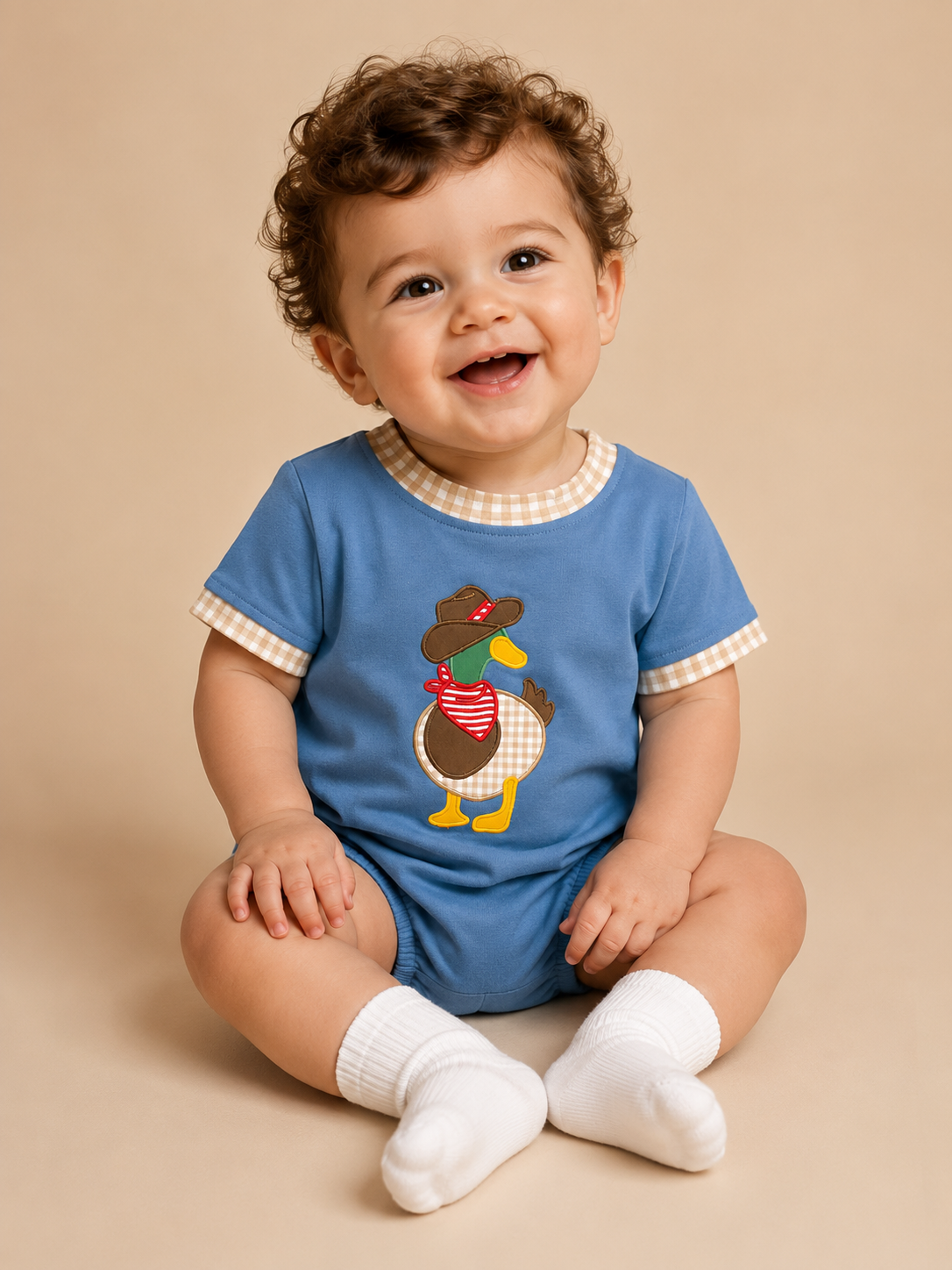 A smiling baby with curly brown hair sits on a beige studio background wearing a blue short-sleeve romper with a gingham collar and sleeve trim, a duck graphic on the front, and white socks.