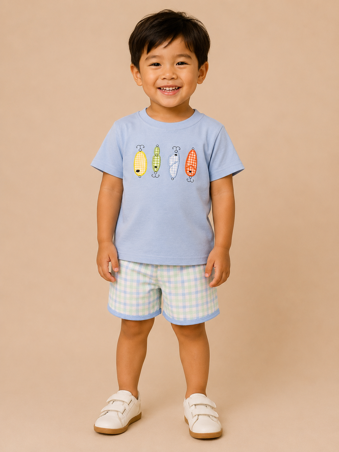 A young child wearing a light blue short-sleeve t-shirt with a colorful fish graphic and light blue and white gingham shorts, standing against a solid beige background.