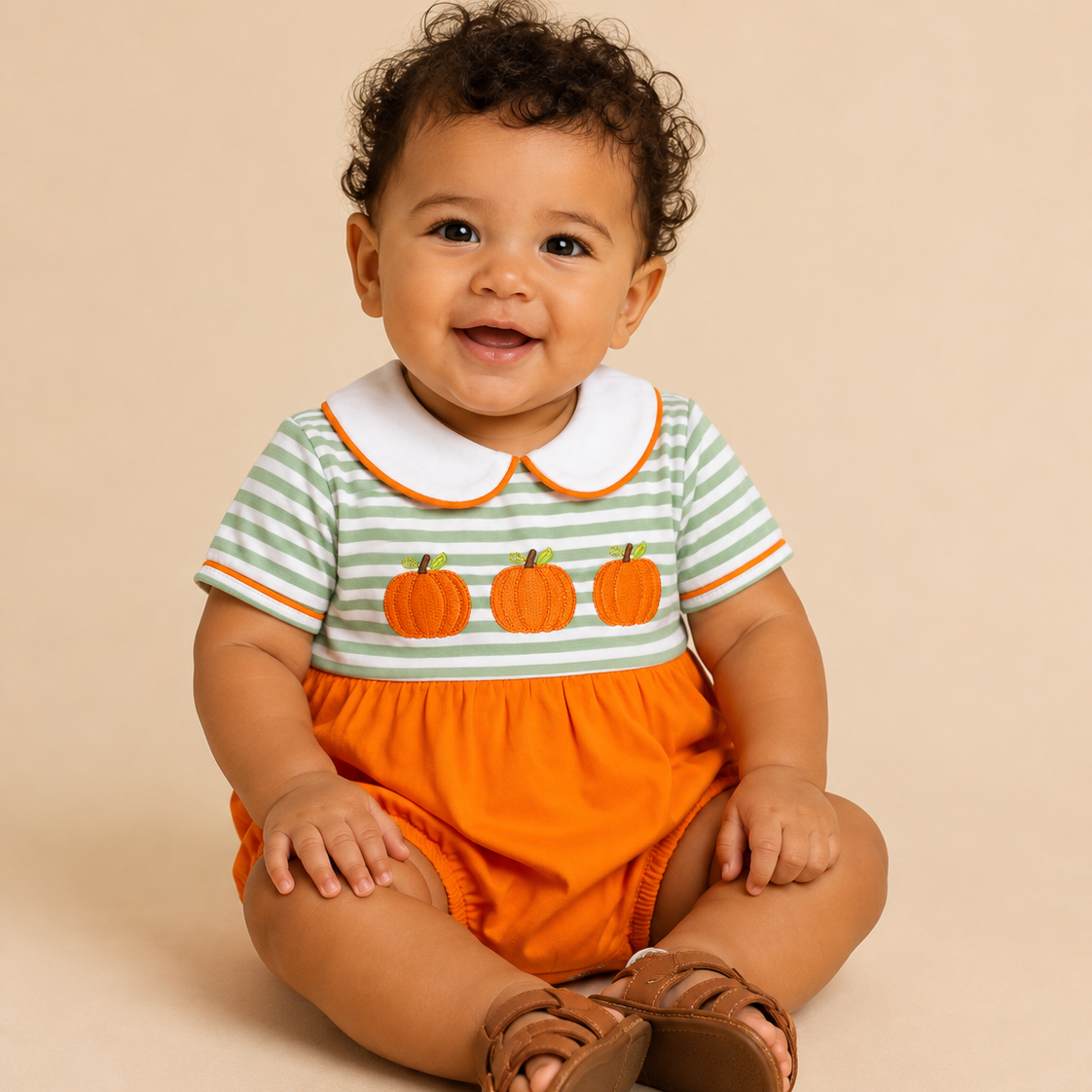 A baby sitting on a beige studio background wearing a short-sleeve romper with green and white stripes, three orange pumpkin appliques, a white Peter Pan collar, and bright orange shorts.