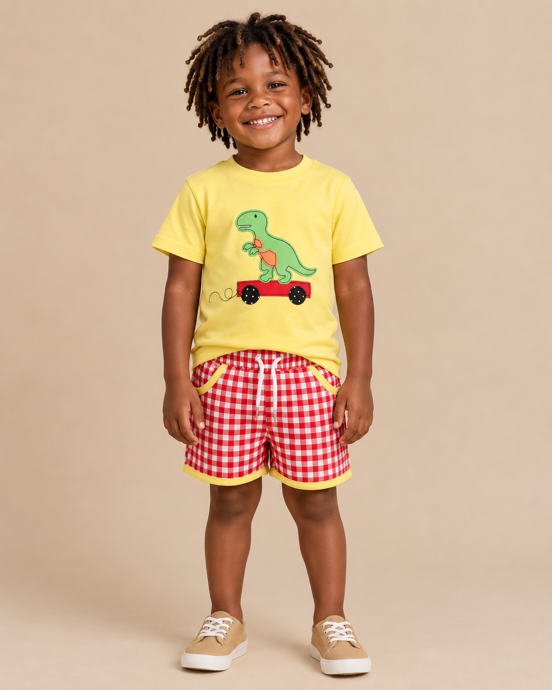 Toddler boy wearing a yellow shirt with a dinosaur on the front and red gingham shorts with yellow trim.