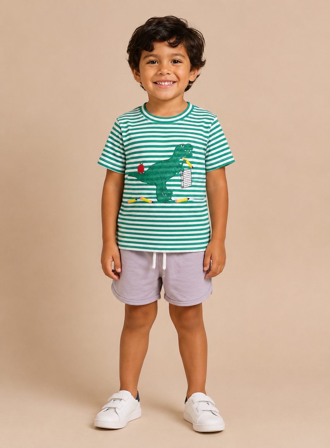 A young boy stands against a solid beige studio background wearing a green and white striped t-shirt with a dinosaur graphic, light purple shorts, and white sneakers.