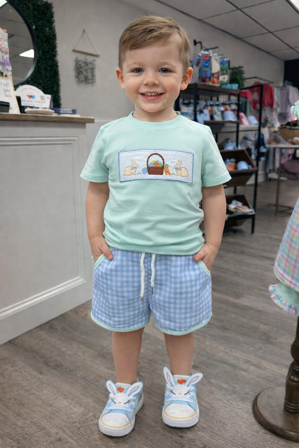 Child wearing a light blue shirt with an Easter graphic design and gingham shorts in a store setting.