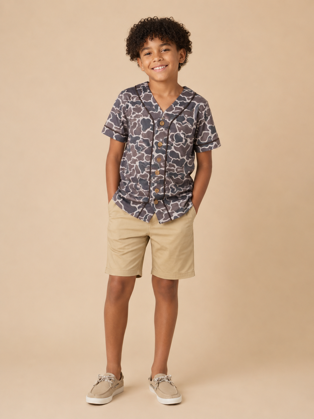 A young boy stands against a solid beige studio background wearing a short-sleeve button-up shirt with a dark gray and white abstract pattern, khaki shorts, and beige sneakers.