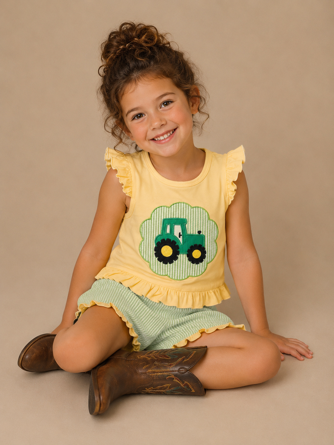 Young girl wearing a yellow top with a green tractor design and green shorts, sitting on a beige background.