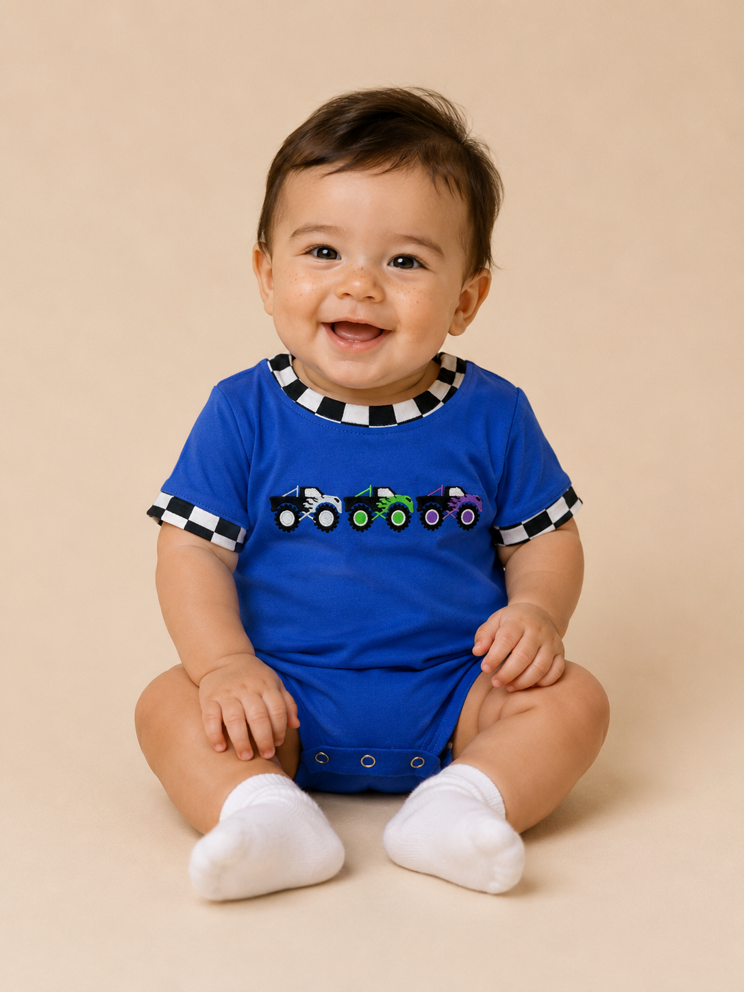 Infant boy wearing a blue bubble with black and white checkered trim, monster trucks embroidered on the front.
