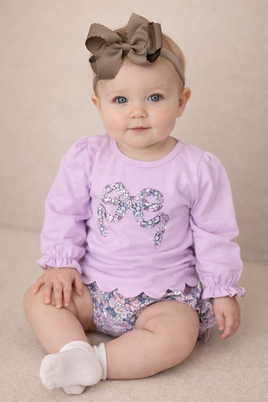 Baby girl sitting down, wearing a lilac top with a floral bow in the middle.