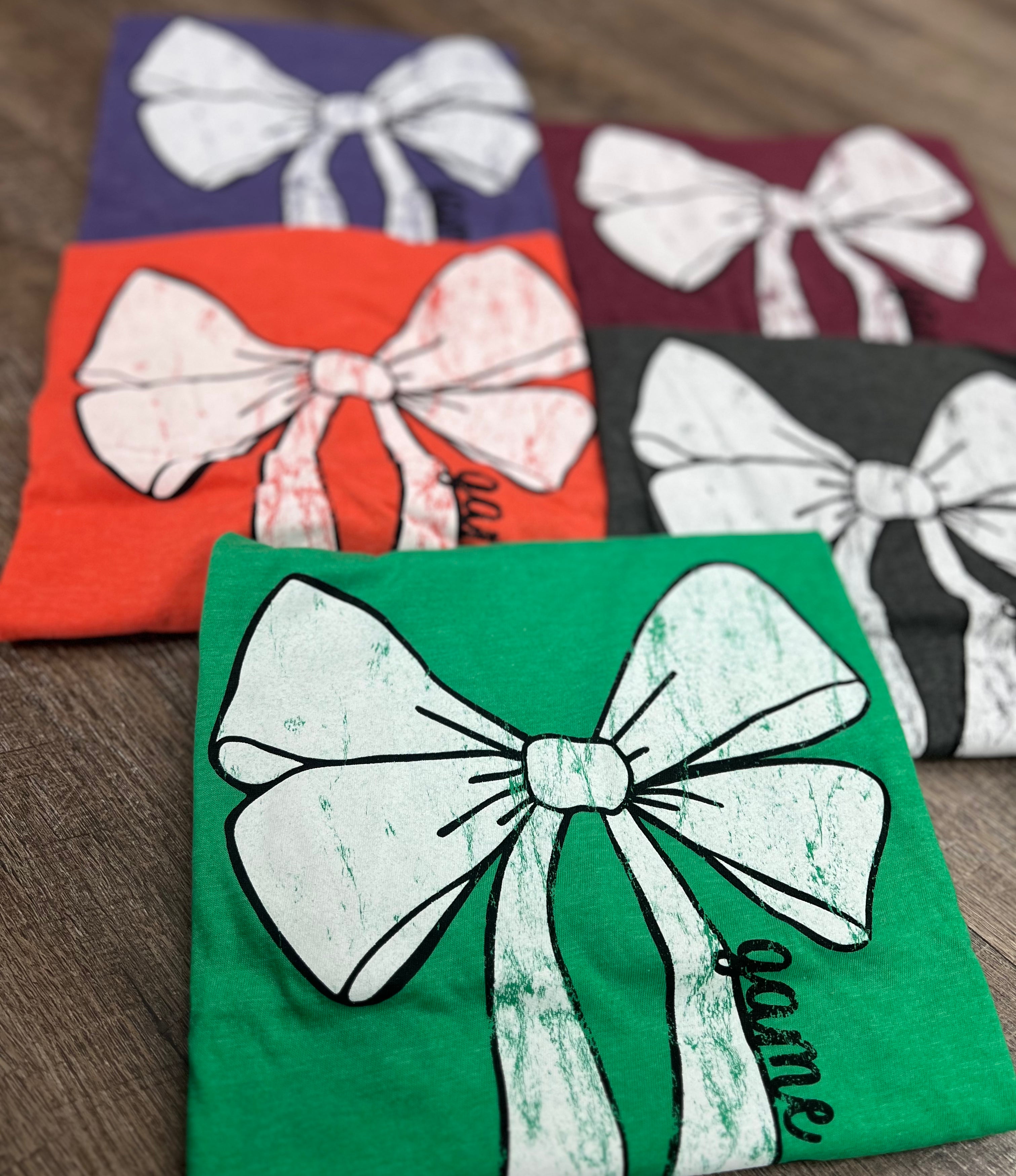 'Game Day' t-shirts with large distressed white bow design. Displayed in five colors: green, charcoal gray, purple, orange, and maroon