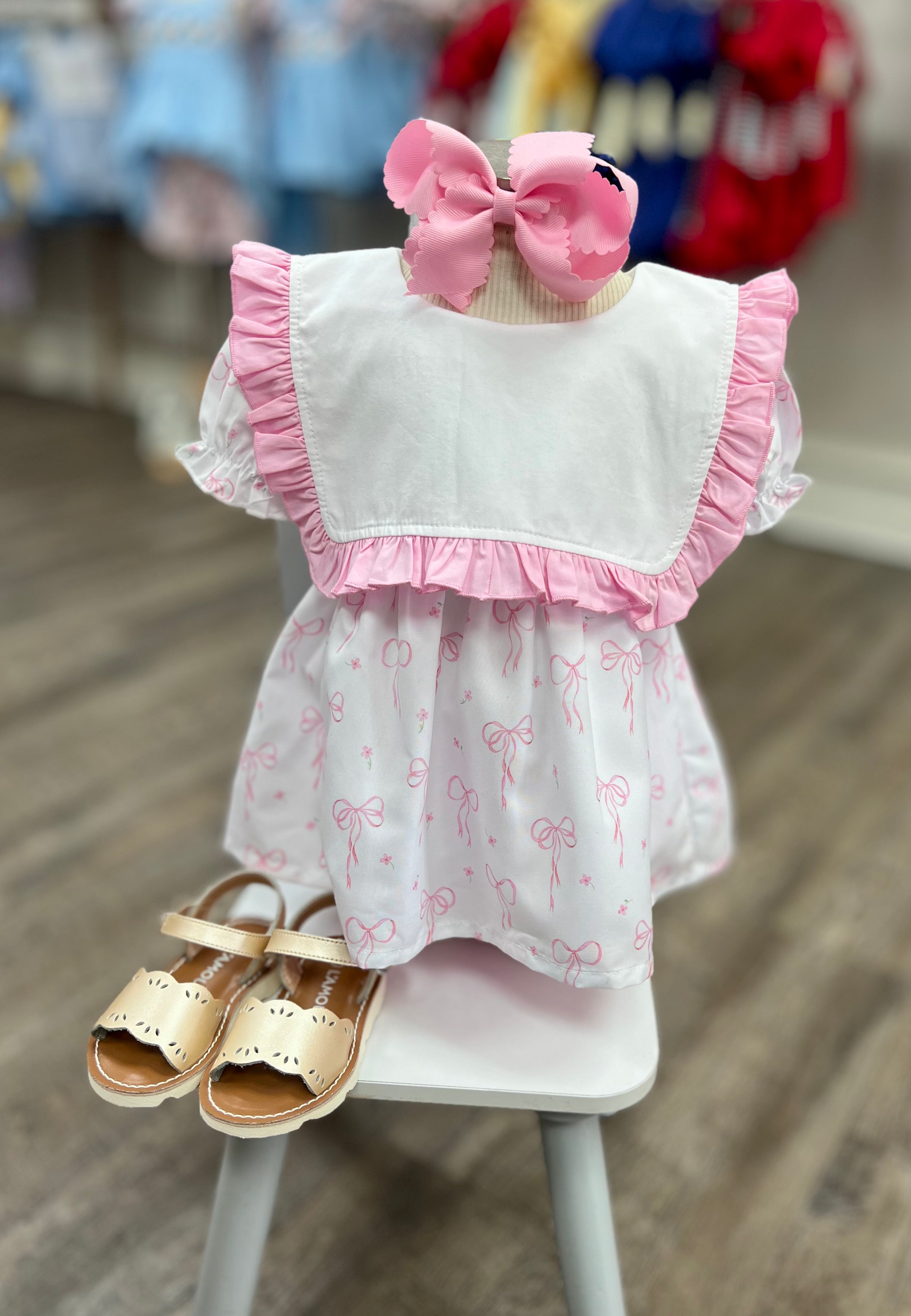 Children's outfit with a white top and pink ruffles, paired with bow-patterned shorts and sandals on a chair.