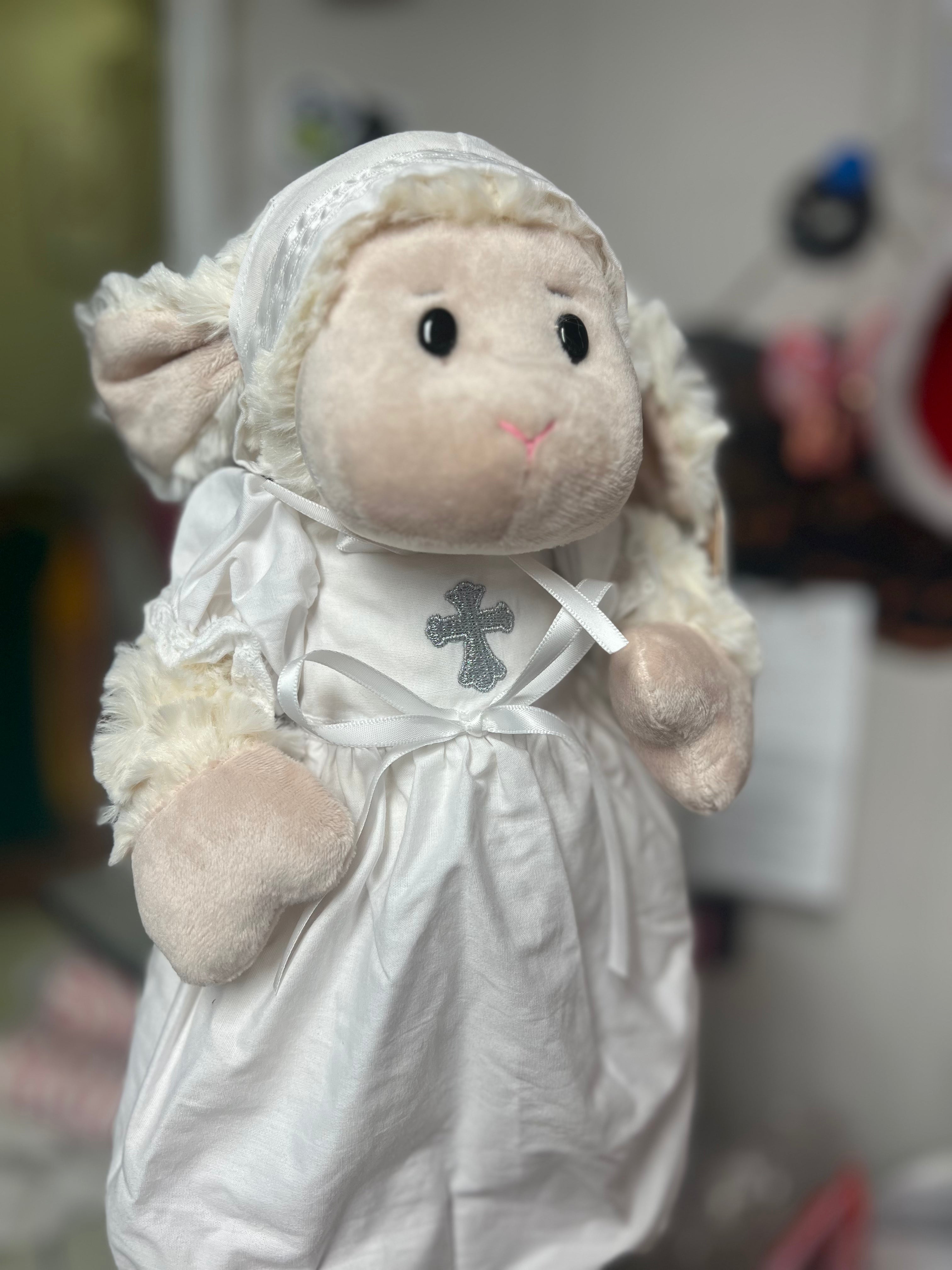Plush lamb toy dressed in a white outfit with a cross, against a blurred background.