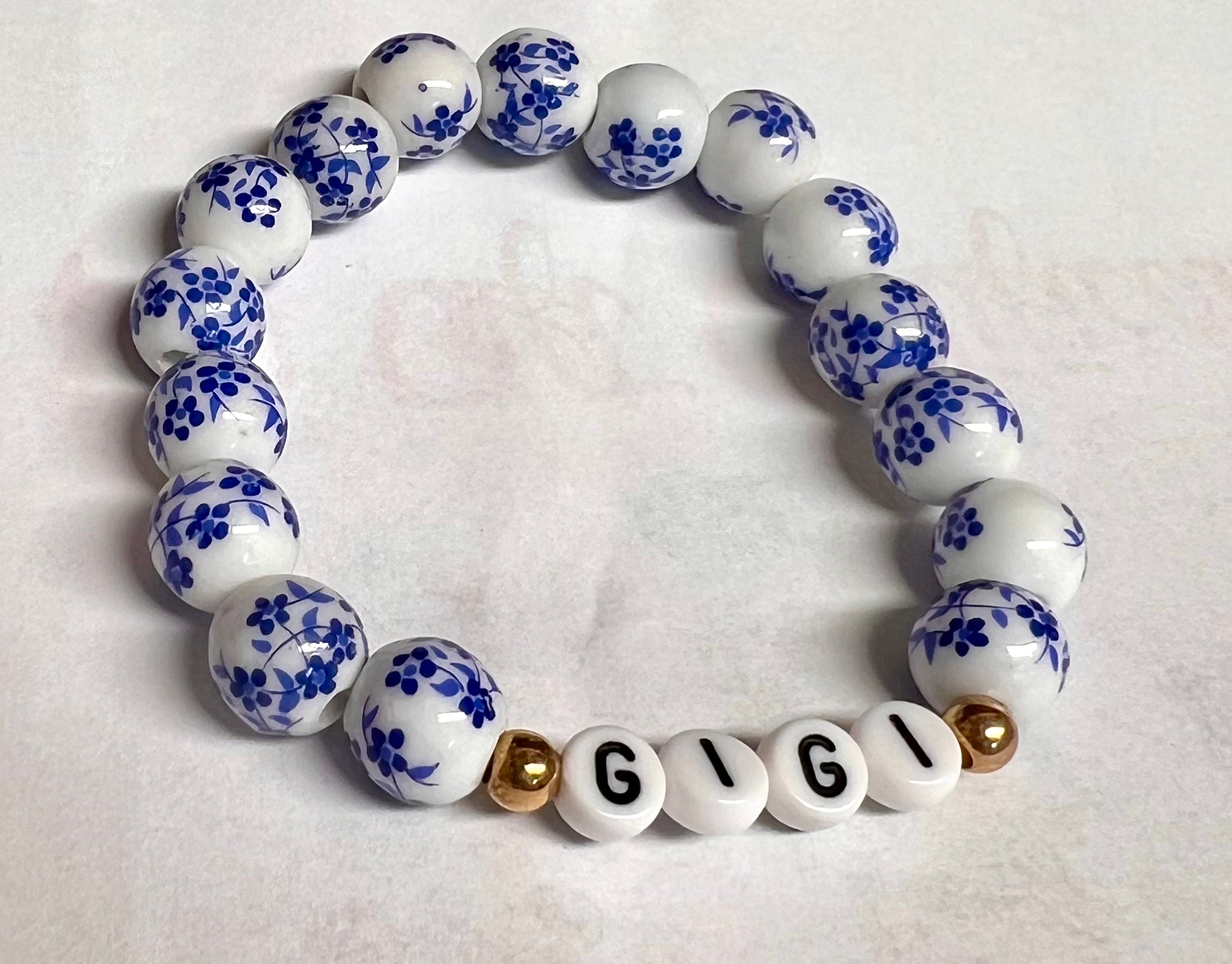 A bracelet with white beads featuring blue floral patterns and gold accents, with the letters 'G I I' in gold-tone letters