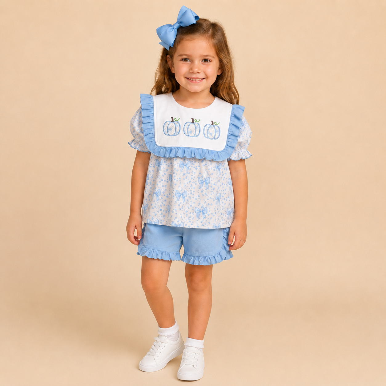A young girl stands against a solid beige studio background wearing a light blue and white outfit consisting of a short-sleeve top with a white bib collar, matching blue ruffled shorts, white socks, and white sneakers. She also has a light blue bow in her hair.
