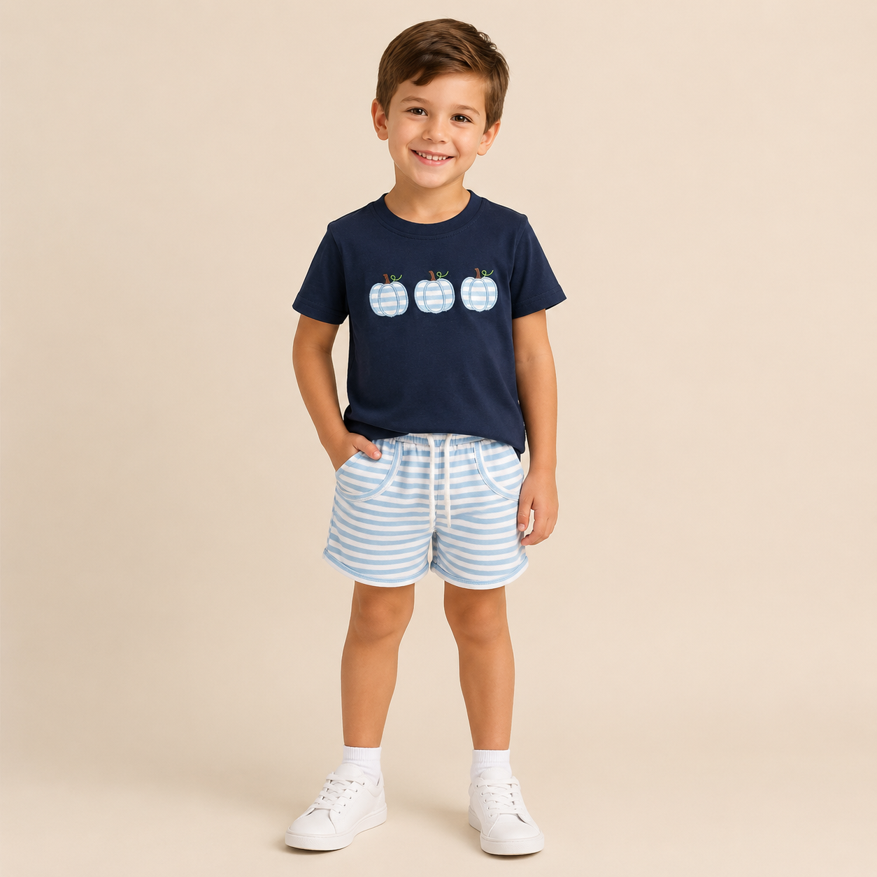 A young boy stands against a solid beige studio background wearing a navy blue t-shirt with three light blue and white gingham patterned pumpkins on the chest, paired with light blue and white striped shorts, white socks, and white sneakers.
