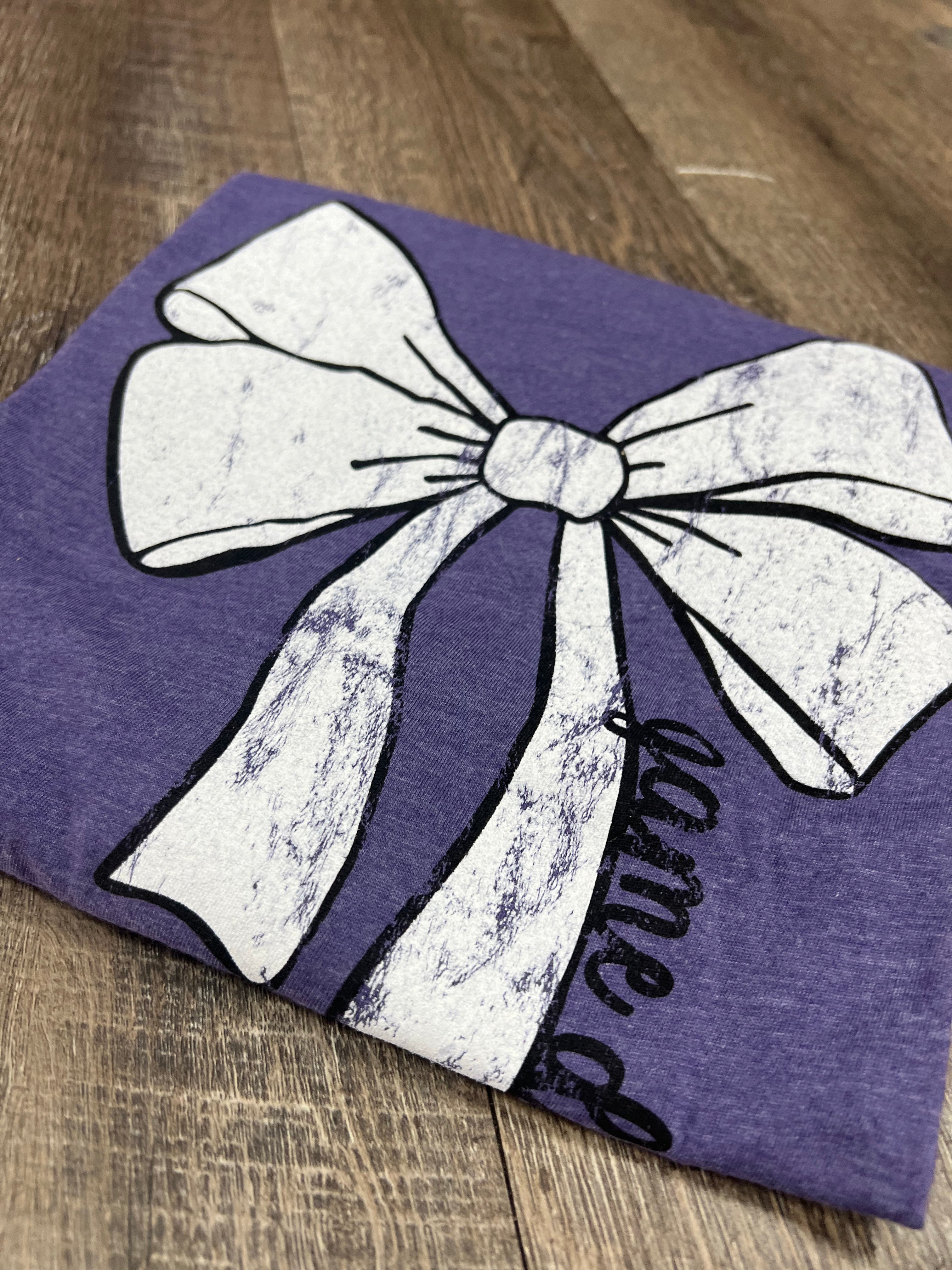 purple t-shirt with white bow outlined in black with game time written on the side of the bow