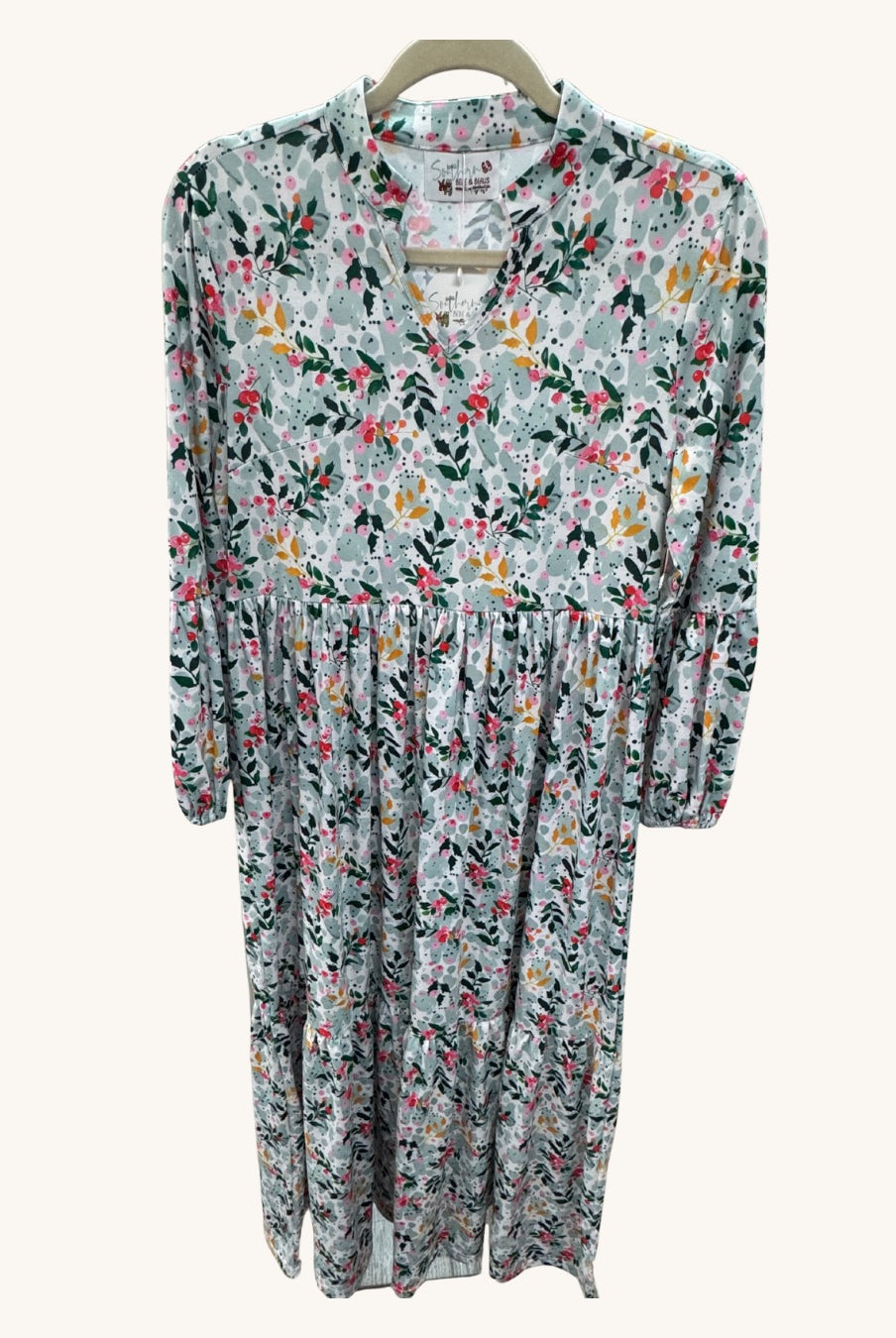 Long floral dress with a light background featuring pink, yellow, and green botanical print, V-neckline, long sleeves, and gathered waist detail.