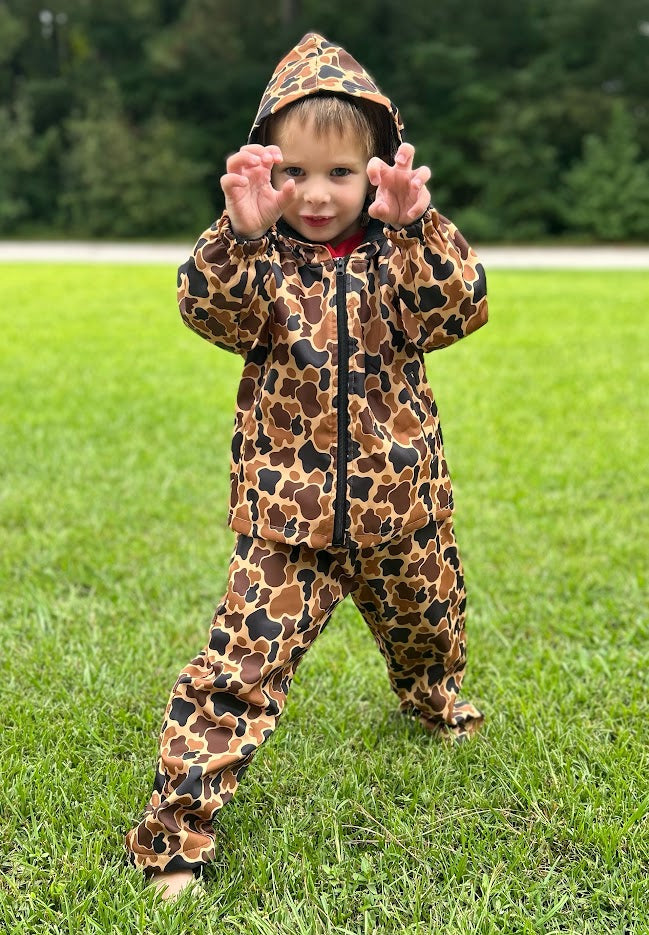 Child wearing a camouflage print raincoat and pants standing on grass.