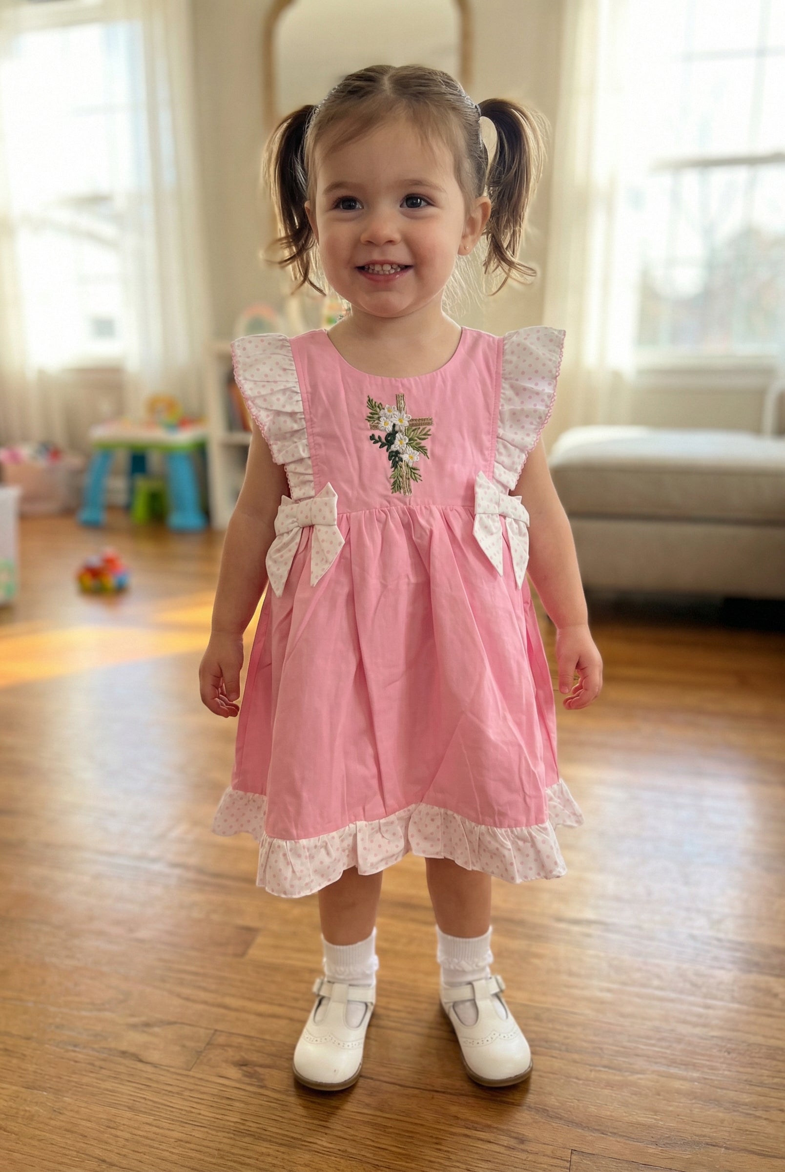 Young girl in a pink dress standing on a wooden floor in a home setting.