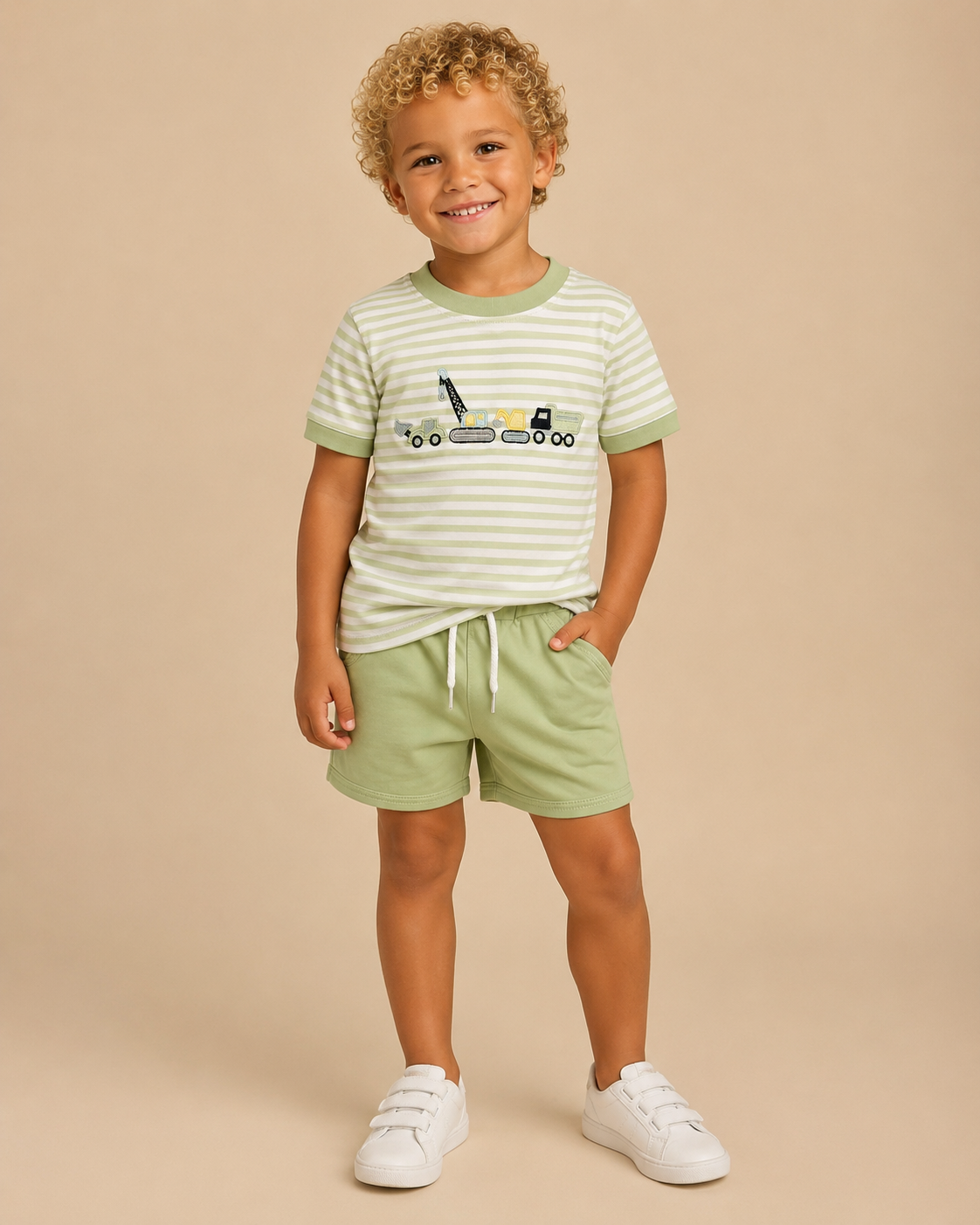 A young child modeling a light green and white striped t-shirt with a construction vehicle graphic, paired with light green shorts and white sneakers, against a solid beige background.
