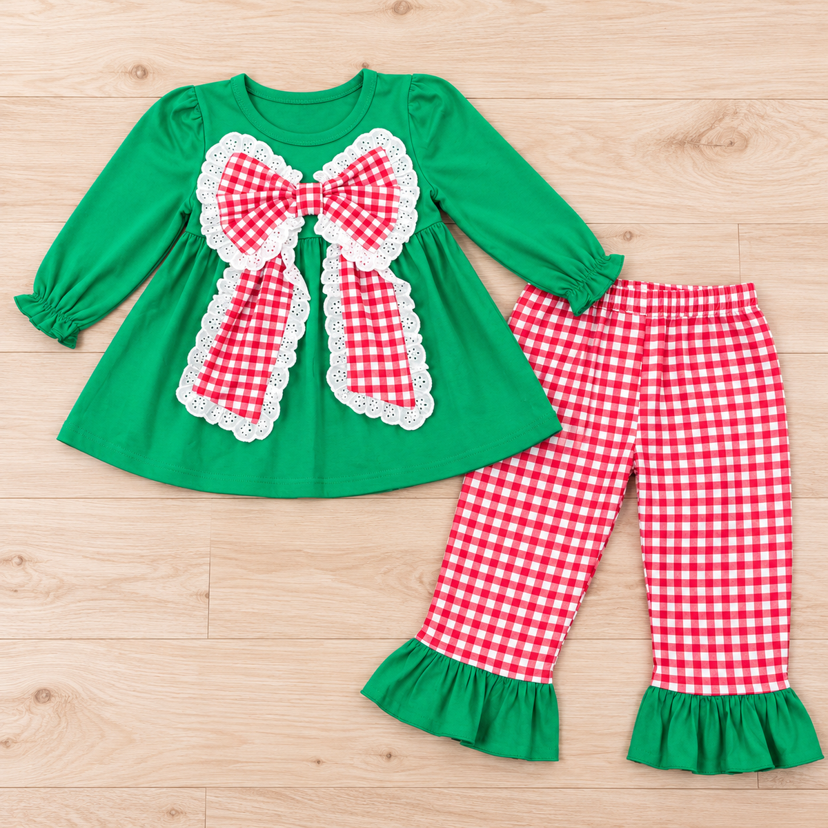 Green girls’ long-sleeve dress-style top with a red gingham bow and white eyelet trim beside red-and-white gingham pants with green ruffle hems.