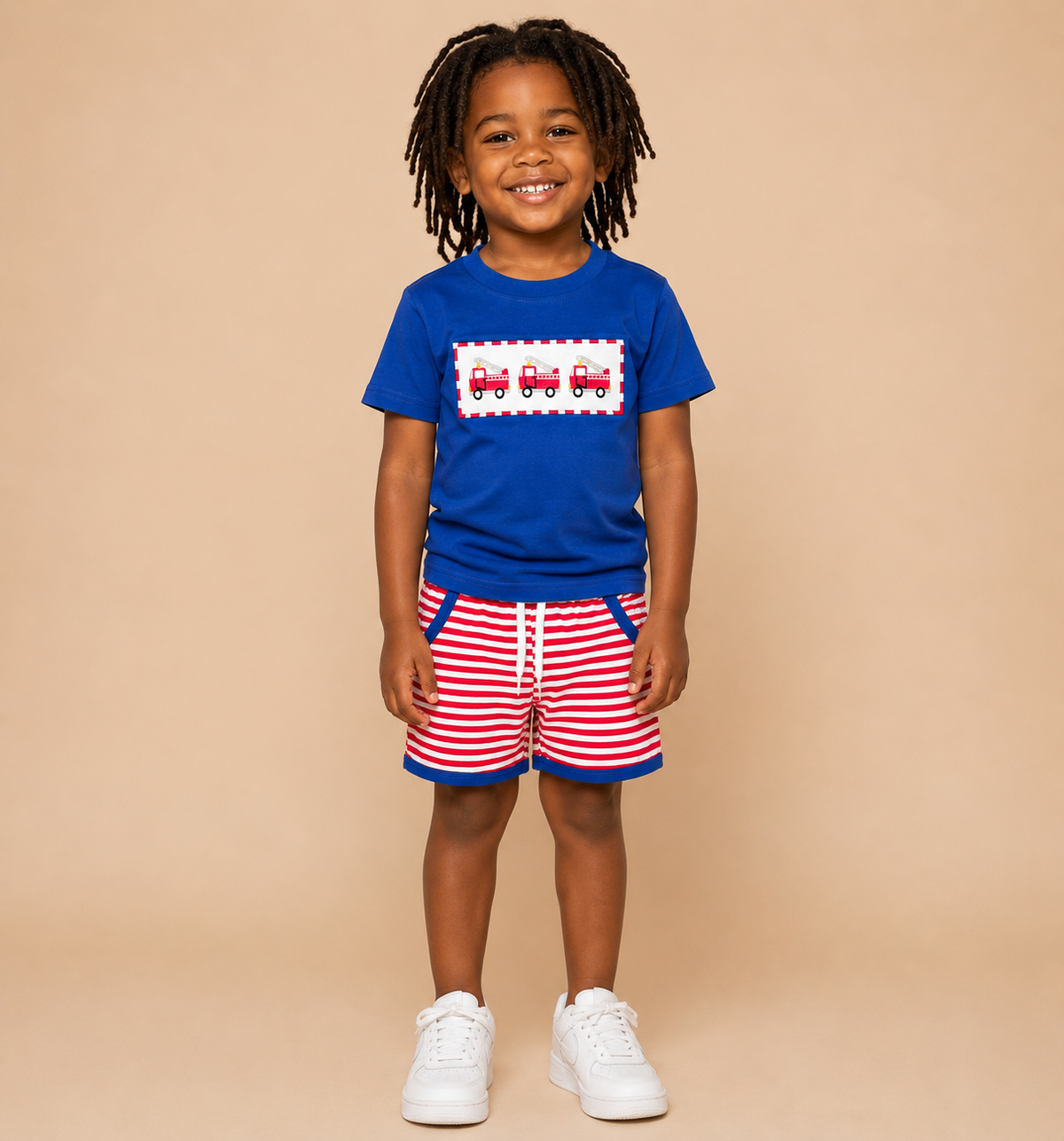 A young child with dreadlocks stands against a solid beige background wearing a royal blue t-shirt with a red and white train graphic, red and white striped shorts, and white sneakers.