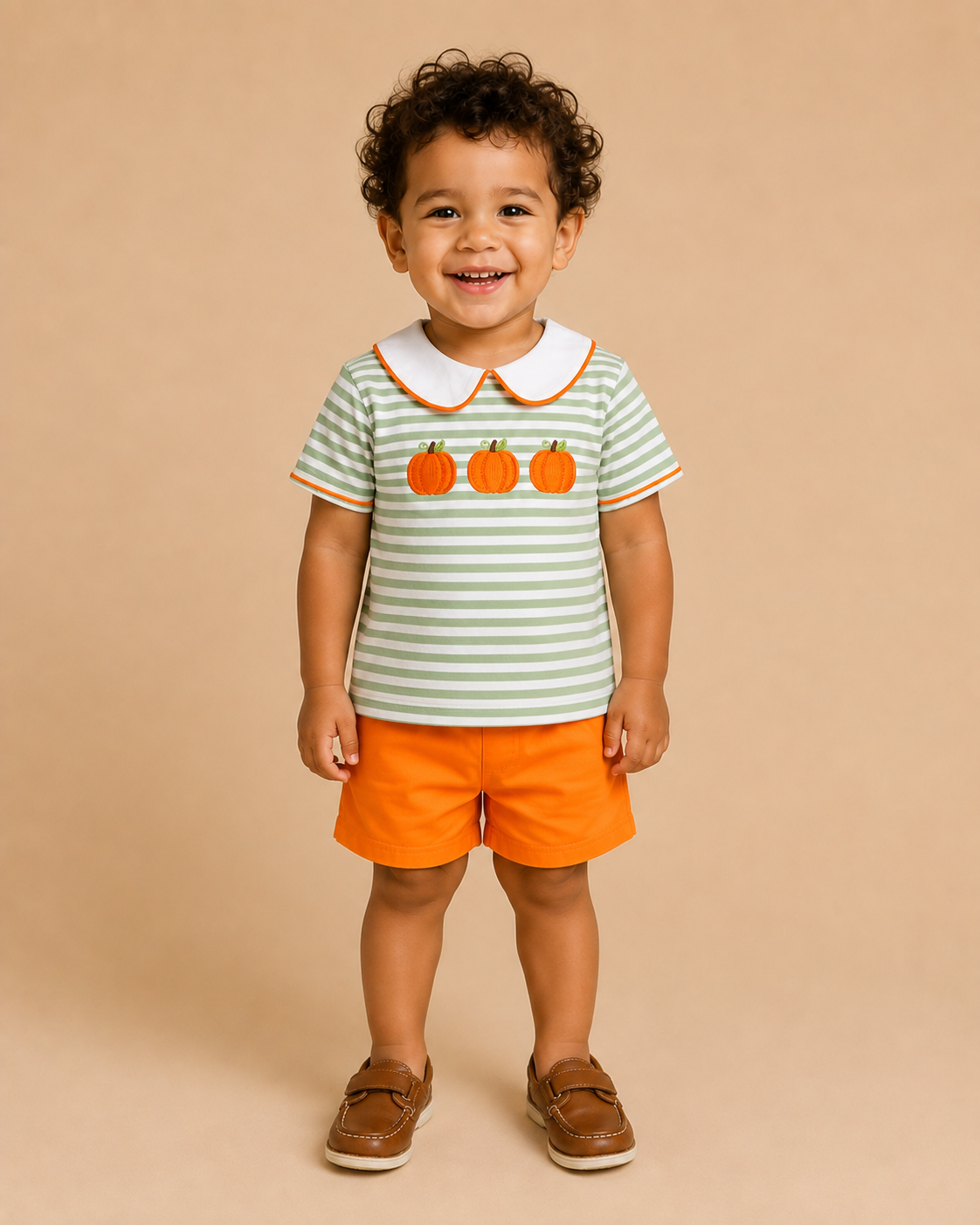 A toddler standing against a solid beige studio background, wearing a short-sleeve polo shirt with green and white horizontal stripes, three orange pumpkins on the chest, a white collar, and bright orange shorts.