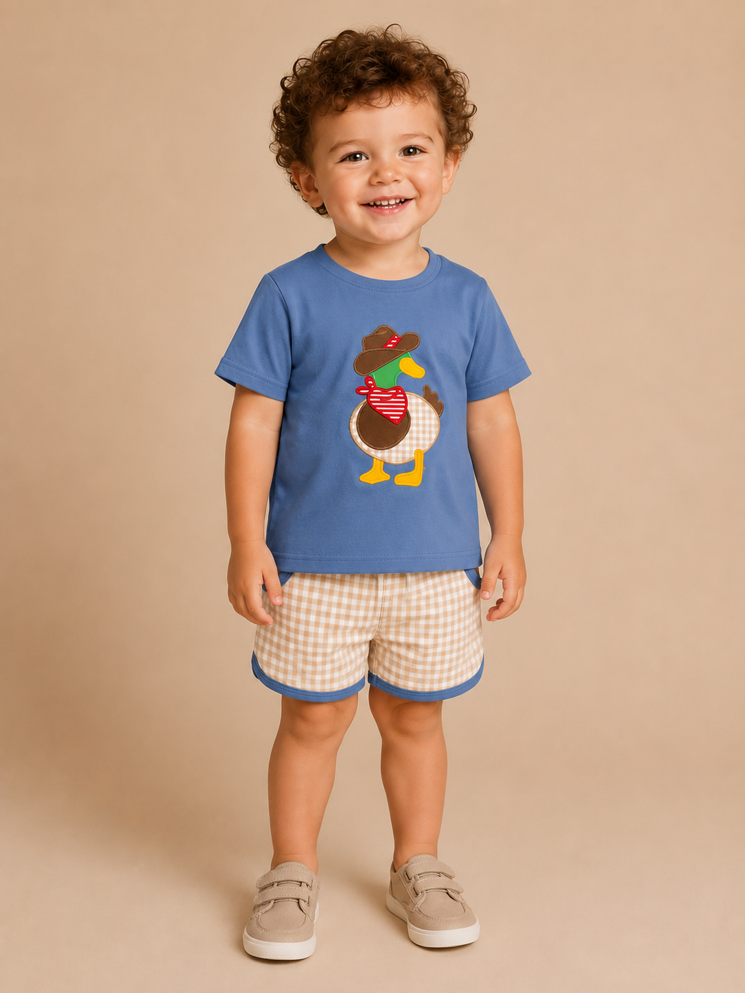 A smiling toddler stands against a warm beige studio background wearing a blue short-sleeve T-shirt with a duck graphic, beige-and-white gingham shorts with blue trim, and beige slip-on shoes.