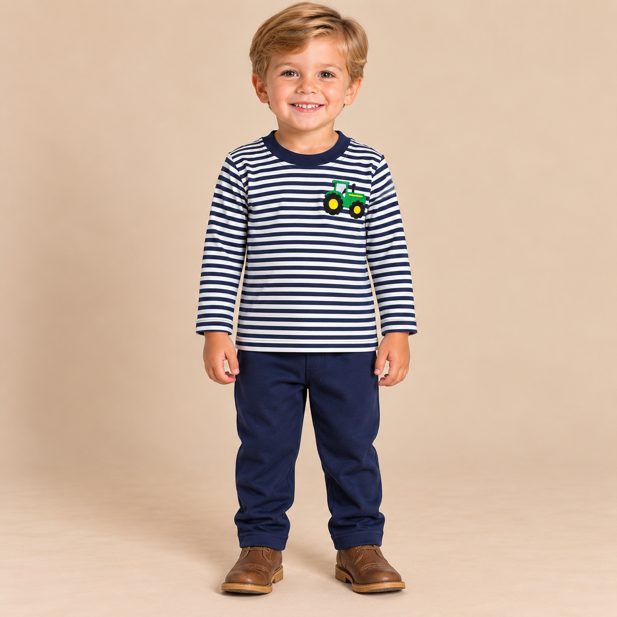 Toddler boy wearing navy and white long sleeve shirt with a tractor wearing navy pants beige background.