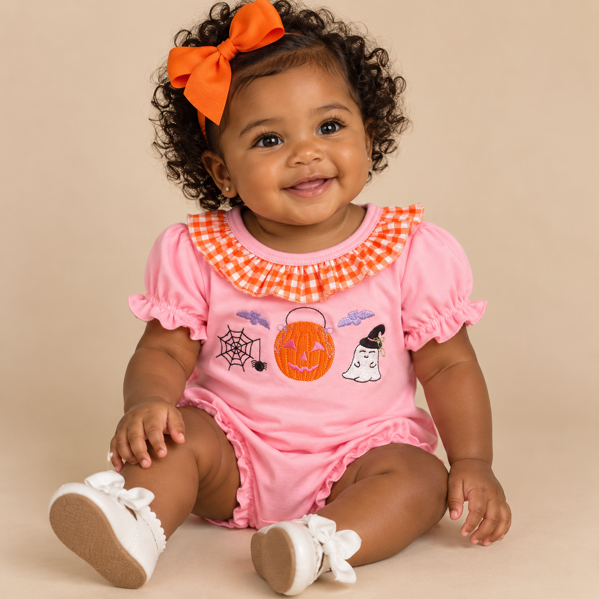 A baby girl wearing a pink short-sleeve bubblewith an orange and white gingham collar, decorated with a pumpkin, spiderweb, and ghost graphic. She is wearing an orange bow in her hair and white baby shoes, sitting against a solid beige background.