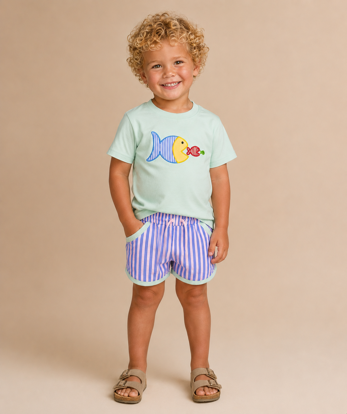 A young child with curly blonde hair stands against a plain beige studio background wearing a mint green t-shirt with a yellow and blue fish graphic.