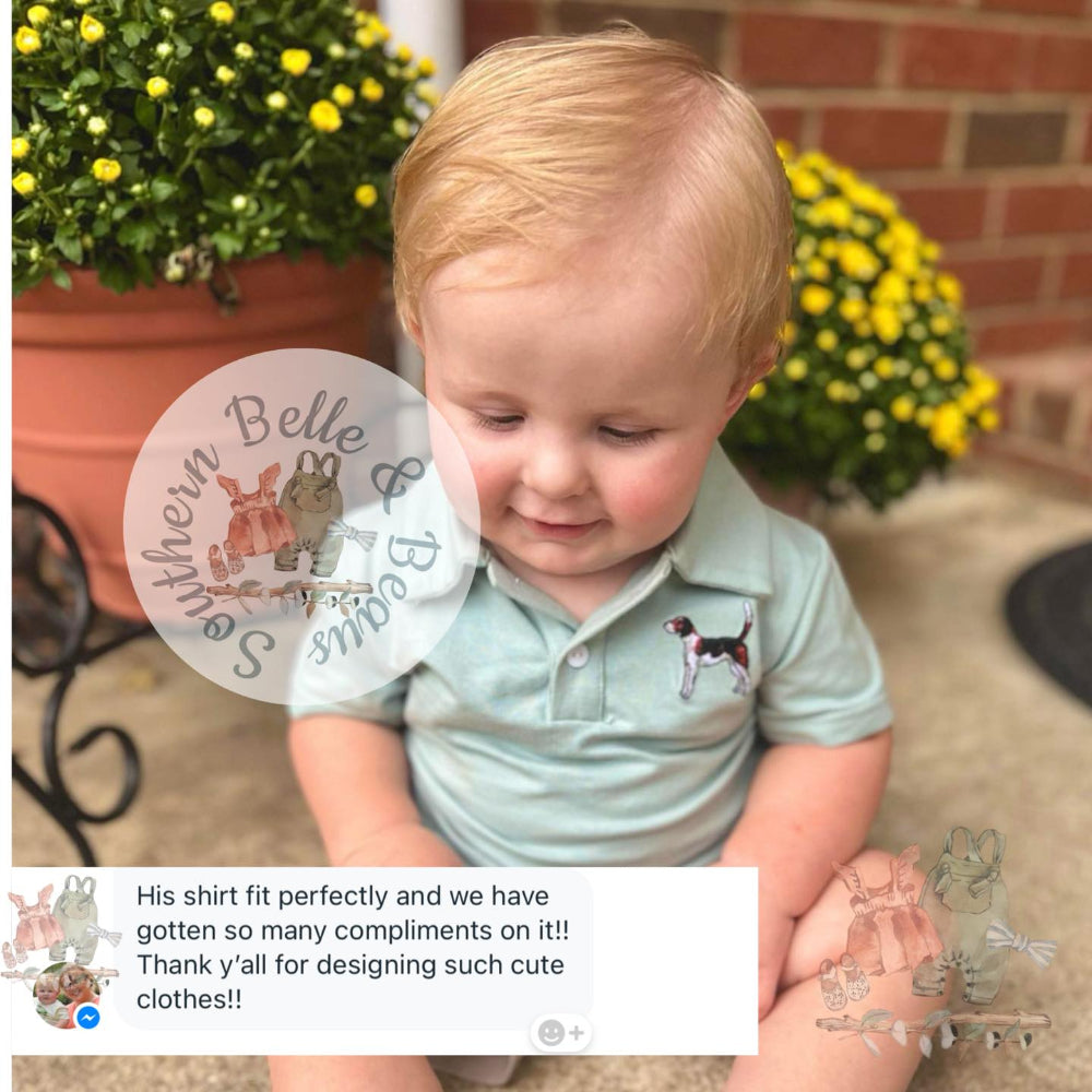 Child wearing a light green shirt with a dog design, sitting outdoors with flowers and a brick wall in the background.