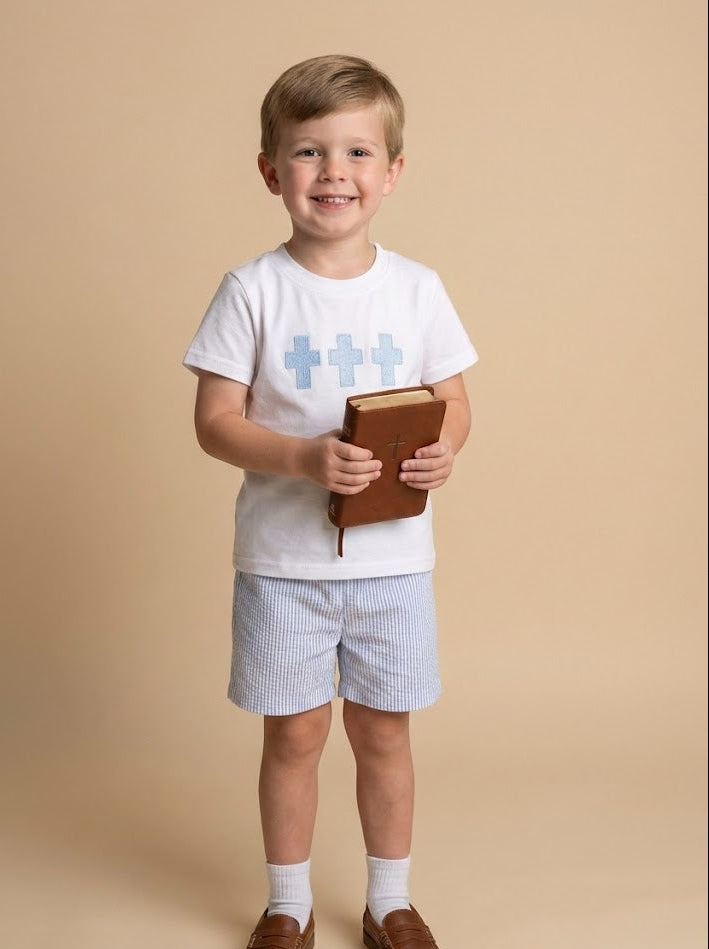 Child wearing a white t-shirt with blue crosses, shorts, and brown shoes, holding a book on a beige background