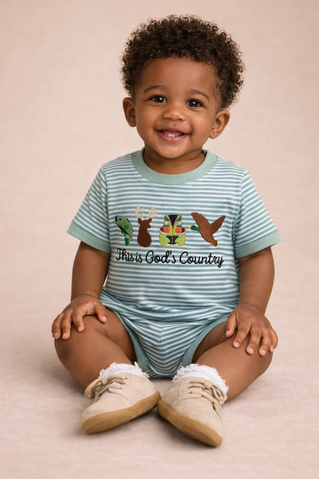 Child wearing a striped onesie with 'This is God's Country' text and animal graphics on a beige background