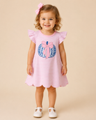 A young toddler girl with curly brown hair wearing a pink and white striped dress with a blue and pink pumpkin applique on the front. She is standing against a solid beige studio background, wearing white Mary Jane shoes and a pink bow in her hair.