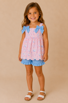 A young girl with long brown hair stands against a solid beige studio background wearing a pink and blue patterned romper with blue bow details on the shoulders and blue ruffled trim at the hem, paired with white sandals.