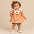 A toddler girl wearing a short-sleeve orange and white gingham dress with three avocado-shaped bib panels featuring pumpkin designs, green ruffled bloomers, a matching orange bow in her hair, and white Mary Jane shoes, standing against a solid beige background.