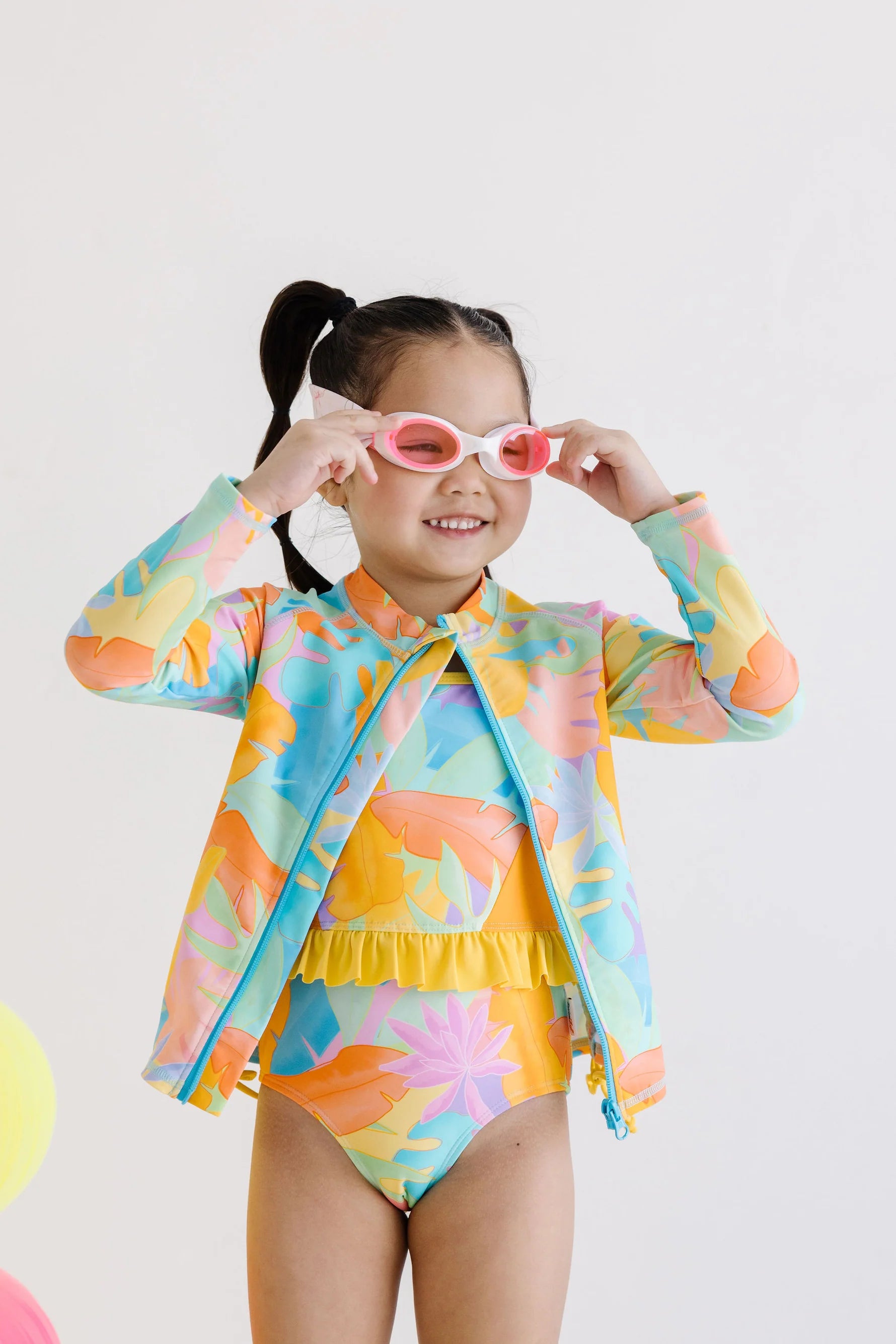 Child wearing a colorful swimsuit and rash guard set with sunglasses on a light background