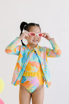 Child wearing a colorful swimsuit and rash guard set with sunglasses on a light background