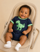 A smiling baby wearing a navy blue short-sleeve bubble with green gingham trim and a green dinosaur graphic, sitting in a beige bouncer chair against a warm beige background.