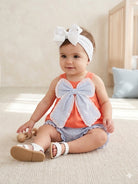 Baby wearing an orange outfit with a large bow sitting on a carpeted floor.