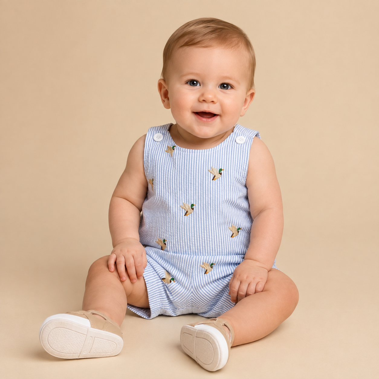 Infant boy wearing a searsucker jon jon with small mallard ducks embroidered on a beige background.