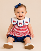A baby girl sitting and smiling, wearing a red and white striped dress with a white bib featuring red fire truck illustrations, navy blue ruffled shorts, a navy blue bow in her hair, and white shoes.