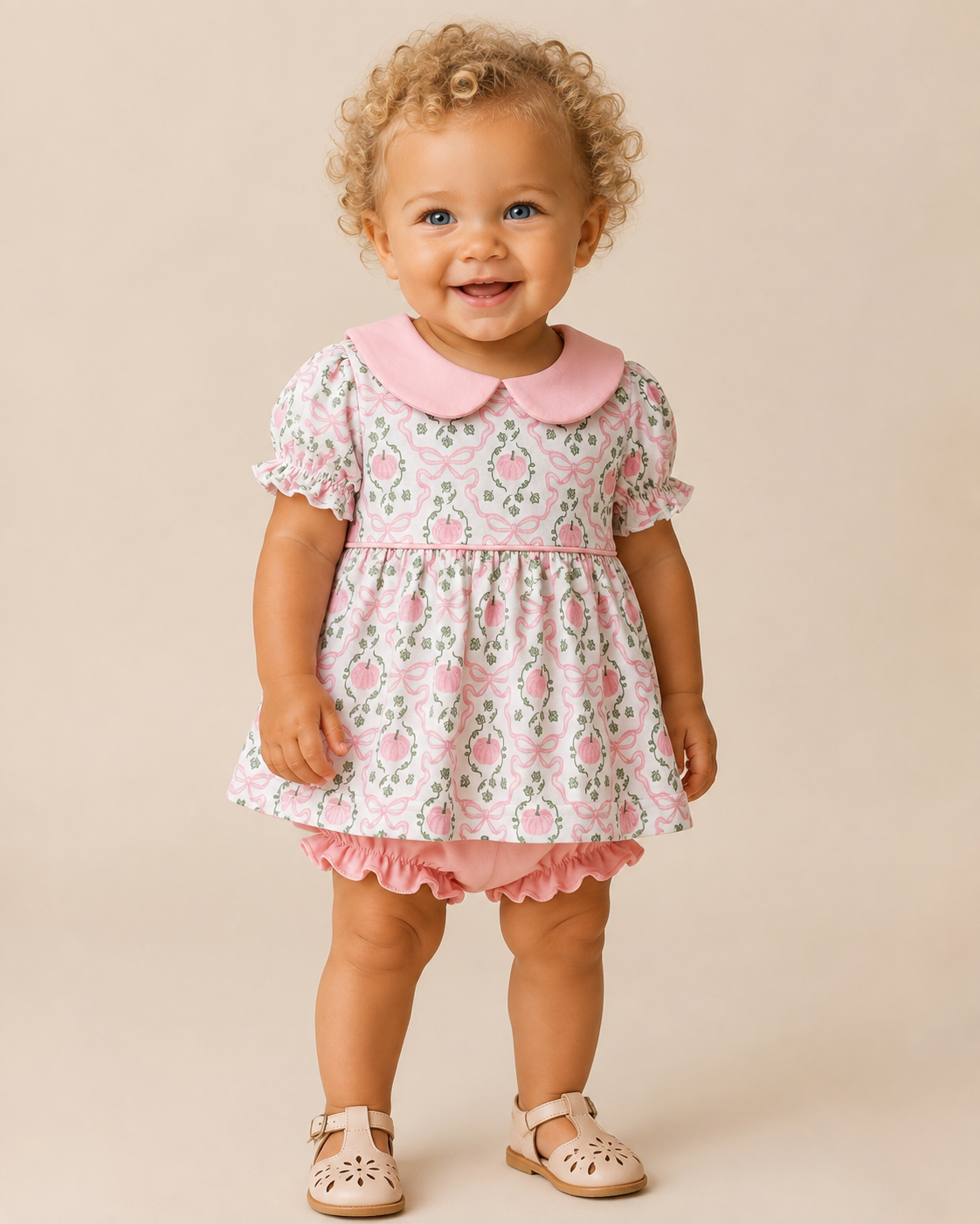 A toddler with curly blonde hair stands against a warm beige studio background wearing a short-sleeve pink dress with a pumpkin & bow print, a pink Peter Pan collar, ruffled trim, and pink booties.