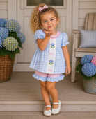 Young girl in a blue checkered dress with floral design, standing on a porch with hydrangeas in the background.