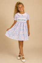Young girl wearing a dress with 'Jesus Loves Me' text on a beige background